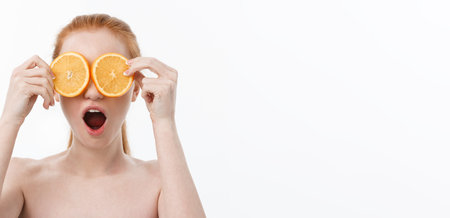 Portrait of a cheerful young girl holding two slices of an orange at her face over white wall backgroundの写真素材