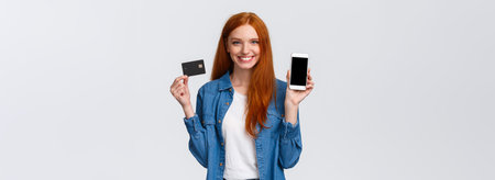 Waist-up portrait confident female with red long hair, blue eyes, holding credit card and showing smartphone display, easy banking service, payment for bills internet, smiling satisfiedの写真素材