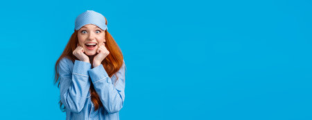 Excited and impressed, amused redhead girl listening fascinating story during sleepover friends house, wearing pyjama and sleep mask, lean on hands and stare camera interested, blue backgroundの写真素材