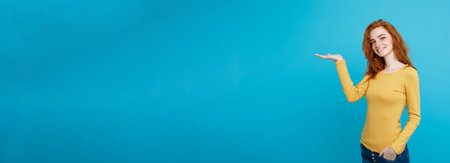 Fun and People Concept - Headshot Portrait of happy ginger red hair girl with presenting hand away and happy expression. Pastel blue background. Copy Space.の写真素材