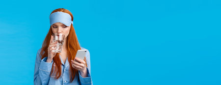 Focused and telephone addicated redhead caucasian woman with glamour sleep mask, nightwear, drinking water from glass and peeking at smartphone screen, chatting over blue backgroundの写真素材