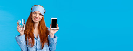 Waist-up portrait elegant and glamour redhead female in nightwear, sleep mask, holding glass water and smiling, holding smartphone, showing mobile app on screen, blue backgroundの写真素材