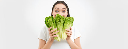 Portrait of cute young woman shows her face with cabbage, likes vegetables, eats healthy nutritious diet food, holds lettuce, white backgroundの写真素材