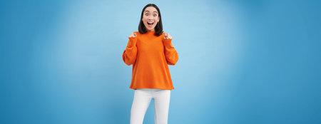 Cheerful asian girl champion, rooting for smth, raising hands up joyful, smiling and celebrating,...