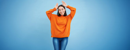 Funny and cute young asian woman shows bunny ears gesture on top of her head and smiling, dancing...