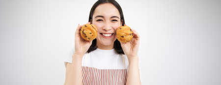 Image of korean woman enjoys eating baked pastry, showing two tasty cupcakes near face and smiles, white backgroundの写真素材