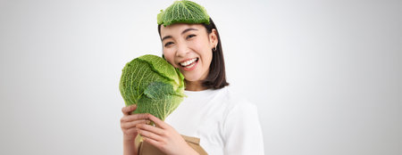 Close up portrait of healthy smiling asian woman, showing cabbage, green letture, with leaf on head, isolated on white backgroundの写真素材