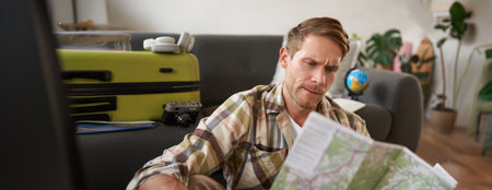 Portrait of happy young man, tourist with suitcase, sits in living room and looks at map, chooses holiday destination, picks route for travellingの写真素材