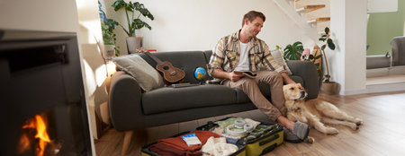 Smiling handsome guy and his dog, sitting in living room, packing suitcase for summer holiday, planning a vacation, holding notebookの写真素材