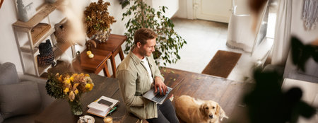 Vertical shot of handsome young man with a dog, leaning on table, holding laptop in hands, looking at screen, checking emails at work. Businessman at co-working space with his golden retrieverの写真素材