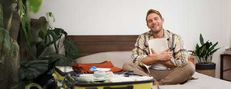 Portrait of charismatic, handsome man, tourist looking at notebook, getting ready for journey, packing suitcase, checking list of items on journalの写真素材