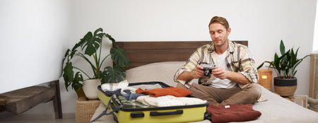 Portrait of happy, smiling young man sitting on bed, unpacking his suitcase after a holiday, tourist looking at his photo album on cameraの写真素材