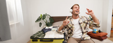 Portrait of cheerful handsome man, tourist going on vacation, dancing and listening to music in headphones while packing suitcase with clothes for holiday trip abroadの写真素材