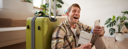 Happy man with suitcase, holding passport and using smartphone, going on holidayの写真素材