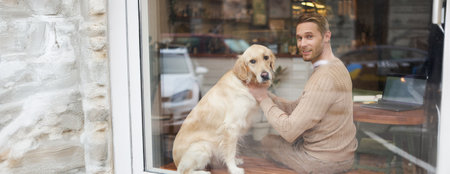 Outdoor shot of a pet-friendly cafe where man sits near the window with his fluffy golden retriever and smiles at camera. Coffee shop visitor with a dogの写真素材