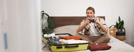 Cheerful young man takes a picture on digital camera, sits on bed, goes on holiday, packs clothes for vacation, tourist excited before going on trip, travellingの写真素材