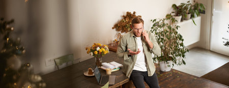 Portrait of man, business manager, talking on mobile phone with concerned, nervous face expression, having a serious conversation, sitting on tableの写真素材