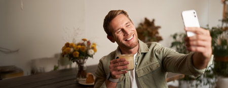 Close up portrait of happy, charismatic young blond man, taking selfie on smartphone, posing with cup of coffee, enjoys his drink in cafe, recommending a place to his followers onlineの写真素材