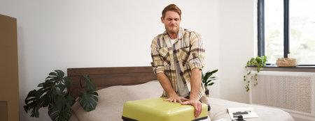 Portrait of young man, tourist packs his bag, closing full suitcase, trying to push the luggage inside to zip the bag, sitting on bed in bedroomの写真素材