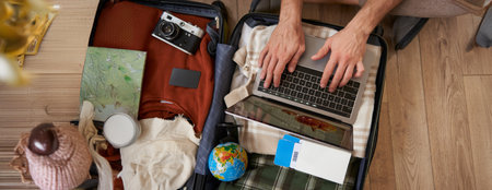 Close up of tourist, hands typing on laptop, opened suitcase with clothes for holiday, man packs luggageの写真素材