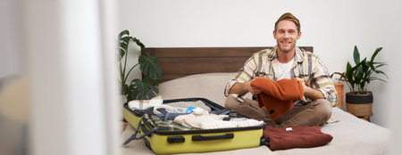 Portrait of happy young man unpacking suitcase after his journey, sitting on bed, getting clothes out of bagの写真素材