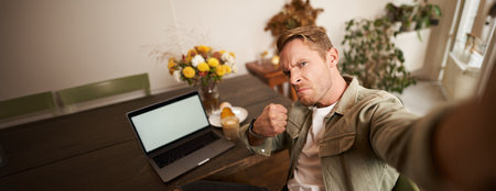 Portrait of handsome man making face for selfie photo, taking picture on smartphone app in cafe, sitting near laptopの写真素材