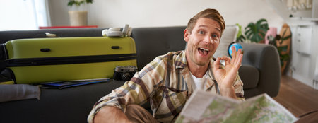 Smiling handsome tourist, man with road travel map, sits on floor near suitcase, shows okay, ok sign, recommending a tourの写真素材