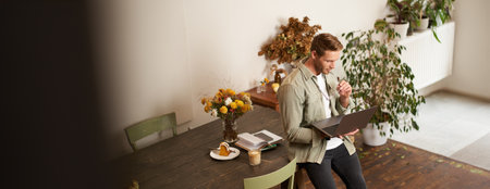 Portrait of young manager, man sits on table, holds laptop, looks concentrated at screen, reading project information, checking work data, working in office or co-working spaceの写真素材