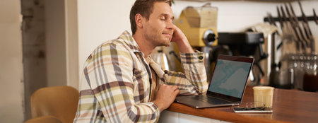 Close up portrait of handsome man, sitting at counter and looking at laptop screen with pleased smile, drinking coffee, browsing on the internet, watching something on computerの写真素材