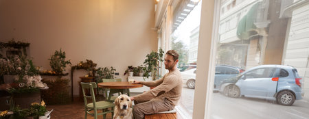Vertical shot of young man, owner of golden retriever, sitting in cafe with and stroking his dog under the table, drinking coffee in a coffeeshopの写真素材