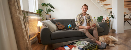 Portrait of young man with check list of items in hands, goes on holiday with his dog, packs suitcase for vacationの写真素材