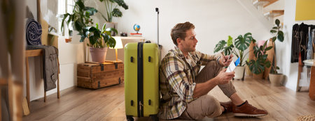 Smiling handsome guy looks at his passport and flight ticket, sits on floor with suitcaseの写真素材