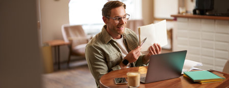 Portrait of handsome adult man showing his exercise to tutor, studying online from coffee shop, sitting in cafe with laptop, pointing at sentence in notebookの写真素材