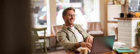 Portrait of man working in cafe on laptop, looking satisfied and pleased with himself, wearing glasses for computer screens, sits back on chair in coffee shopの写真素材