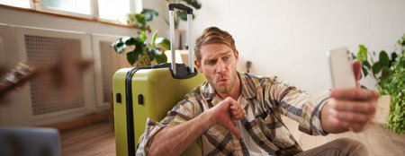 Disappointed tourist, young man giving negative feedback, shows thumbs down, takes photo on smartphone, sits near travel suitcaseの写真素材