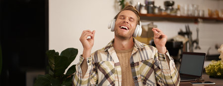 Handsome smiling young man, digital nomad, sitting in co-working open space, working in coffee shop, singing and listening to music in wireless headphonesの写真素材