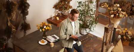 Portrait of handsome young man sits with laptop, looks at screen with concentrated, thoughtful face, working on a project, e-learning at co-working space, drinking coffeeの写真素材