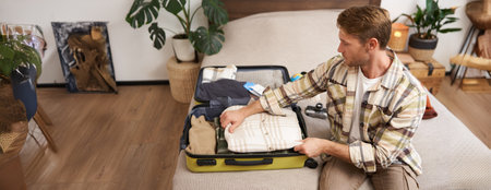 Tourism and travelling concept. Portrait of handsome young man going on holiday, packing his suitcase, checking luggage, sitting on bed and looking at packed thingsの写真素材