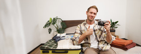 Portrait of handsome man going on vacation, packing suitcase, holding camera, travel photographer going on holidayの写真素材