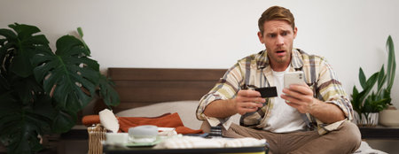 Portrait of worried young man, sitting on bed with suitcase, holding credit card and mobile phone,...
