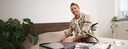 Portrait of young handsome bearded guy, tourist going on holiday, fastening his clothes in suitcase,...
