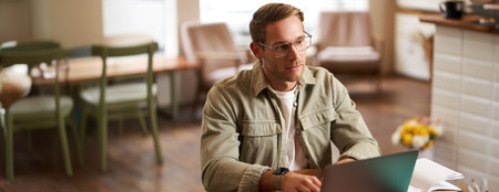 Portrait of young concentrated man in glasses, working from coffee shop, studying, looking...