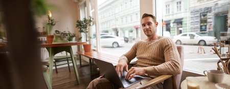 Portrait of young working professional, man sitting in cafe with laptop, drinking coffee and working...