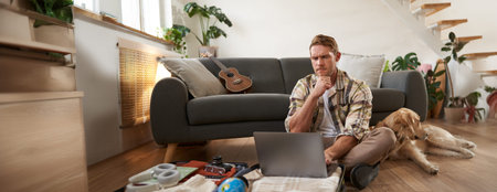 Image of young man, tourist going on holiday, sitting with a dog in living room, looking at laptop,...