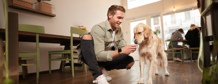 Portrait of smiling handsome man with his dog, sitting on floor in cafe with golden retriever, giving a treatの写真素材