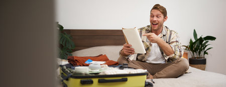 Portrait of charismatic, handsome man, tourist looking at notebook, getting ready for journey, packing suitcase, checking list of items on journalの写真素材