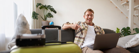 Happy handsome guy, sits with laptop, points finger at screen and laughs, has packed suitcase and camera prepared for holiday, goes on vacationの写真素材