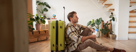 Portrait of smiling handsome tourist, man holding passport and flight tickets, sitting with suitcaseの写真素材