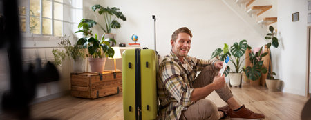 Portrait of handsome young man, tourist sitting with his suitcase, holding passport and flight tickets, going on holidayの写真素材