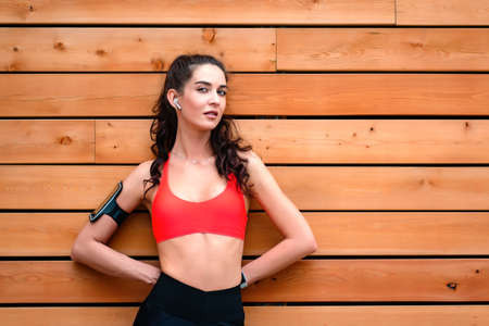Young attractive smiling toned brunette woman with earphones and phone holder on arm outside, wooden background, copy space. Healthy lifestyle concept. Fit beautiful woman portraitの写真素材